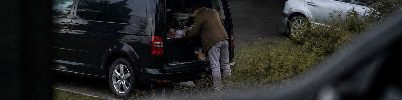 Ein Mann am ge&ouml;ffneten Heck eines Transporters auf einer Parkfl&auml;che, beobachtet aus einem Fahrzeug heraus