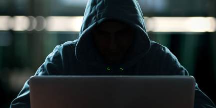 Vermummte Person mit Kaputzenpullover sitzt an einem Laptop