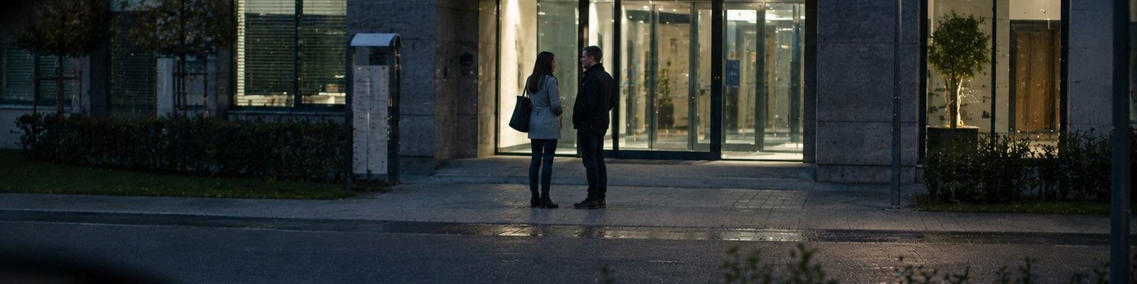 Zwei Personen am Abend vor einem B&uuml;roeingang, beobachtet aus einem Fahrzeug heraus